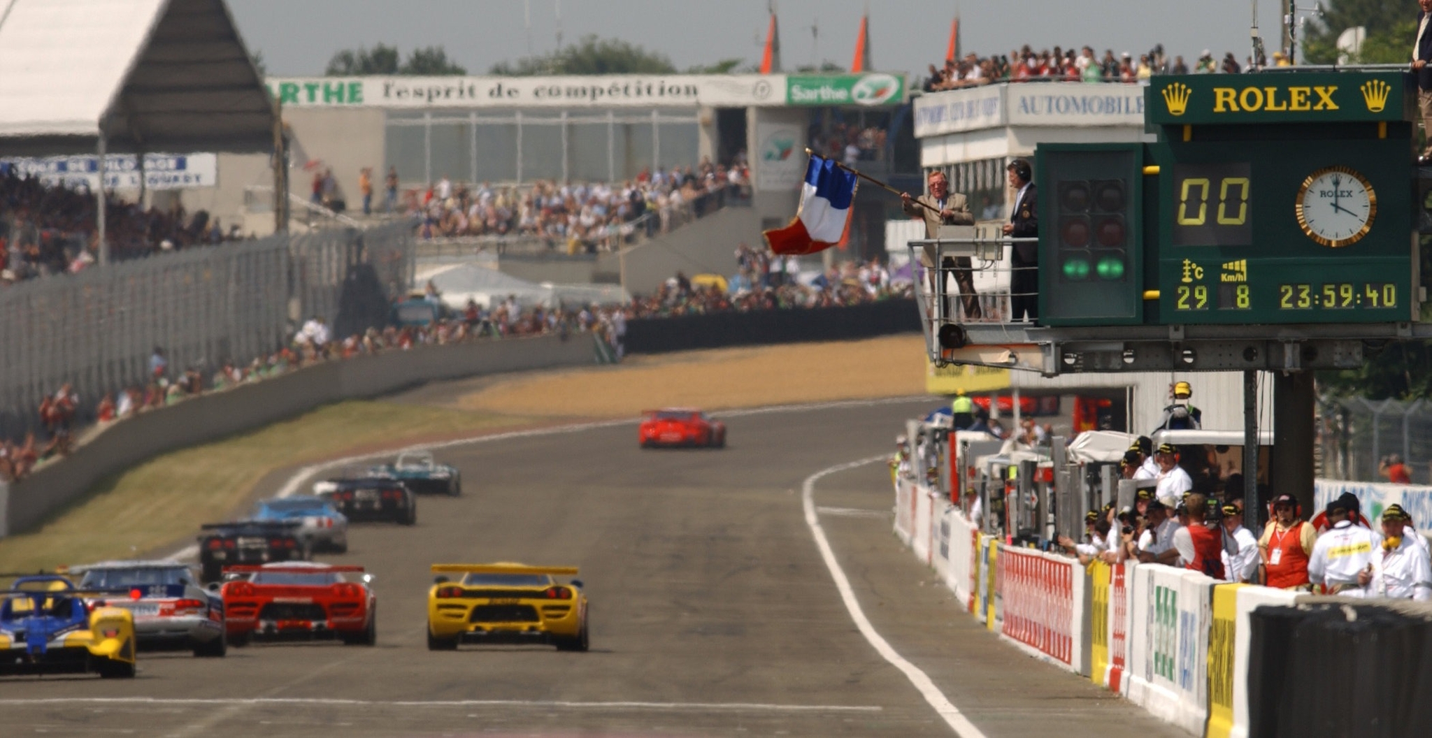 Đoàn xe trên đường đua 24 Hours of Le Mans cạnh bảng giờ Rolex